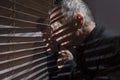 Mature man looking out of a window with blinds casting shadows