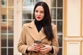 Horizontal image of serious confident businesswoman holding papercup of coffee, looking directly at camera, posing over glass door