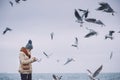 Horizontal image of young beautiful woman feeds seagulls on the sea. Pretty female wearing coat, scarf, hat watching flying Royalty Free Stock Photo