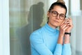 Horizontal indoor image of beautiful blonde young serious woman in office wearing blue turtleneck and eyeglasses, folded hands Royalty Free Stock Photo