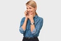 Horizontal indoor image of young pretty blonde woman in denim shirt smiling and talking on smart phone to her friend Royalty Free Stock Photo