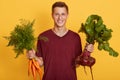 Horizontal indoor picture of cheerful happy adorable young boy holding bunch of carrots and beet, looking directly at camera, Royalty Free Stock Photo