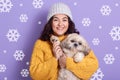 Horizontal indoor picture of cheerful smiling curly haired woman standing isolated over lilac background, holding Maltese dog in