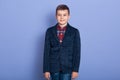 Horizontal indoor shot of positive sweet child standing isolated over lilac background, wearing jeans, plaid shirt and dark jacket Royalty Free Stock Photo