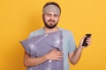 Horizontal indoor shot of sleepy miserable man wearing sleeping mask and t shirt, holding pilow and smartphone, waking up, putting