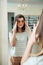 Horizontal indoor shot of trendy modern woman in casual outfit standing near mirror in optician store, trying on stylish