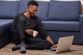 Horizontal indoorm shot of dark skinned male posing on floor near blue sofa, watches funny video on laptop computer or having