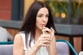 Horizontal outdoor shot of thoughtful attractive woman enjoys day off, poses in outdoor cafe, drinks milkshake, dressed in white b