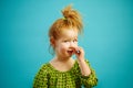 Horizontal photo of cute redheaded girl picking her nose on isolated blue background, has a beautiful face.