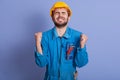 Horizontal picture of exhausted construction worker clenching his fists, raising hands, closing eyes, being tired of work, wearing