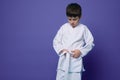 Horizontal portrait of a confident teenage boy aikido wrestler tying white belt of his kimono,  over violet background Royalty Free Stock Photo