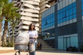 Horizontal portrait. Happy young mother pushing a pram with baby, walking in the city against high rise office buildings Royalty Free Stock Photo