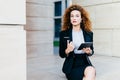 Horizontal portrait of pretty confident businesswoman dressed formally, holding her pocket book with pen and tablet computer, bein