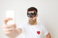 Horizontal portrait of serious young male model with thick beard and mustache wears batman black mask, casual white t Royalty Free Stock Photo