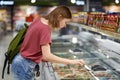 Horizontal shot of adorable teenage female going to buy frozen vegetables, looks into friedge while strolls in grocer`s shop, dres Royalty Free Stock Photo