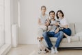 Horizontal shot of affectionate family pose together on couch in empty spacious room with white walls, their favourite dog sits on