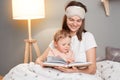 Horizontal shot of attractive happy smiling mother reading a book for her baby daughter while posing in bed, child looking at Royalty Free Stock Photo