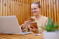 Horizontal shot of attractive winsome young adult woman with bun hairstyle wearing beige sweater working on laptop, holding paper Royalty Free Stock Photo