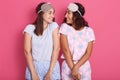 Horizontal shot of attractive young women standing over pink background, posing in sleeping mask and pajamas, looking at