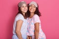 Horizontal shot of beautiful girls posing in pajamas and sleeping mask, girls friends standing issolated over pink background,