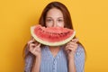 Horizontal shot of beautiful smiling young woman with slice of watermelon, female covering half of face with fruit, lady wearing Royalty Free Stock Photo