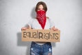 Horizontal shot of beautiful young female belong to feminist move, wears red bandana on face, holds plate with inscription Equal r