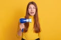 Horizontal shot of beautiful young woman holding blue hairdryer in hand, posing isolated over yellow studio background, having
