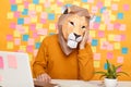 Horizontal shot of bored anonymous man in lion mask wearing orange sweater sitting at workplace with laptop against yellow wall