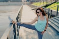 Horizontal shot of of brunette woman wears casual sport clothes stretches legs on fence has athletic body warms up before cardio