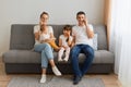 Horizontal shot of busy family sitting on sofa and having conversation on smart phone, looking at camera, kid with smart phone Royalty Free Stock Photo