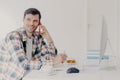 Horizontal shot of busy male freelancer has telephone conversation, dressed in checkered shirt, writes down information, sits at