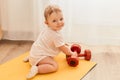 Horizontal shot of charming cute tiny infant kid sitting on yoga mat with dumbbells looking at camera with smile, exploring sport