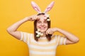 Horizontal shot of cheerful satisfied woman wearing bunny ears holding easter cake pops, looking isolated over yellow background, Royalty Free Stock Photo