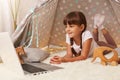 Horizontal shot of cute delighted little girl with braids wearing casual clothing laying on floor on soft carpet in wigwam and Royalty Free Stock Photo