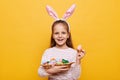 Horizontal shot of cute optimistic little girl wearing rabbit ears holding Easter eggs in wicker basket, showing testicle painted