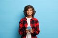 Horizontal shot of cute teenager with curly hair, holds mobile phone, waits for message, smiles gently, dressed in red