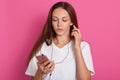 Horizontal shot of dark haired hipster girl listening to audio track with thoughtful facial expression, has ear phones and holding