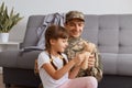 Horizontal shot of delighted father with his kid, Caucasian soldier man wearing camouflage uniform returning home after army, Royalty Free Stock Photo