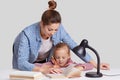 Horizontal shot of experienced young mother leans near her small child, helps to do home assignment, shows what to rewrite in book Royalty Free Stock Photo