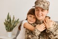 Horizontal shot of extremely happy satisfied woman wearing camouflage uniform and cap posing with her daughter, kid hugging her Royalty Free Stock Photo