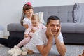 Horizontal shot of extremely happy young adult father lying on floor near sofa with his little kids on back, man keeping hands on Royalty Free Stock Photo