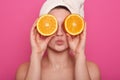 Horizontal shot of funny cheerful young woman holding two halves of orange against her eyes, having white towel on her head, Royalty Free Stock Photo