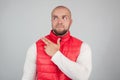 Horizontal shot of handsome bearded male with bald head, dressed in casual red waistcoat, looks curiously asdie, points with index Royalty Free Stock Photo