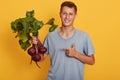 Horizontal shot of handsome model standing isolated over yellow background in gray t shirt, holding raw vegetable in hand, smiling Royalty Free Stock Photo