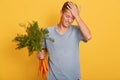 Horizontal shot of handsome young man touching his forehead with hand, holding bunch of carrots in one hand, young guy forgot Royalty Free Stock Photo