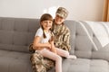 Horizontal shot of happy Caucasian woman wearing camouflage uniform and cap posing with her daughter, sitting on sofa, expressing Royalty Free Stock Photo