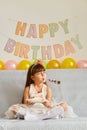 Horizontal shot of happy little girl wearing white dress sitting on sofa with birthday cake and blowing party horn, rejoicing her Royalty Free Stock Photo