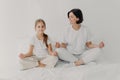 Horizontal shot of happy relaxed brunette mother and daughter pose in lotus pose, try to relax, keep legs crossed, dressed in