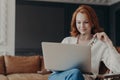 Horizontal shot of happy skilled ginger woman teacher works distantly, checks students works via laptop, holds spectacles, poses