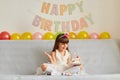 Horizontal shot of happy smiling little girl wearing white dress sitting on sofa with birthday cake and burning candle, rejoicing Royalty Free Stock Photo